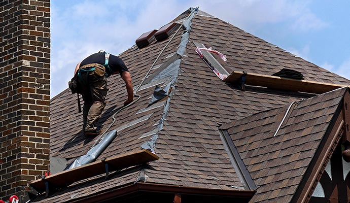 An expert repairing a steep shingle roof using tools and a safety harness near a brick chimney